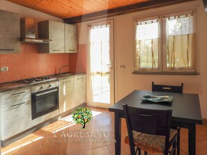 A kitchen with a stove, oven, and sink, a dining table with chairs, and a window with curtains. - L'Agrumeto-Casa Cedro. Porto Tramatzu beach (Teulada)