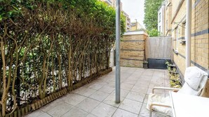 Een terras met betegelde vloer, een metalen stoel, een witte kussen, een hoge metalen paal en een bakstenen muur.
