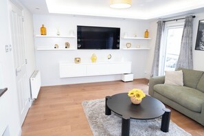Uma sala de estar moderna com televisão de tela plana, prateleiras brancas com objetos decorativos, um sofá verde e uma mesa de centro redonda com um vaso de flores.