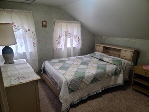 Ein Schlafzimmer mit einem Bett, einem Nachttisch, einer Kommode, einer Lampe und zwei Fenstern mit floralen Vorhängen.