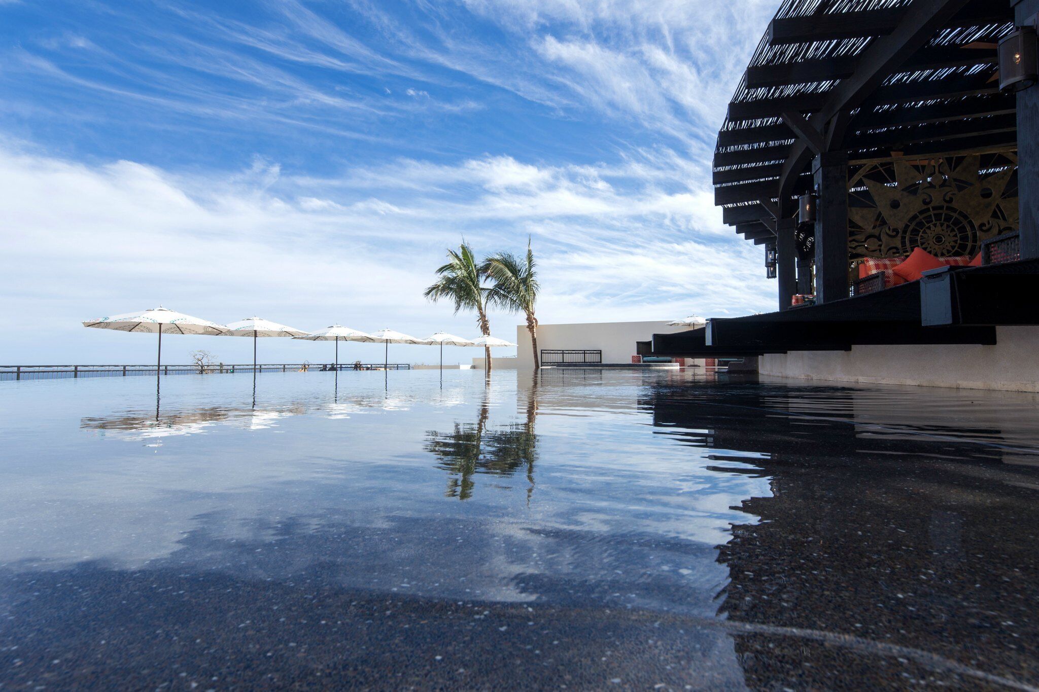 Ein spiegelnder Pool mit Sonnenschirmen, Palmen und einem Gebäude mit ornamentiertem Bau.