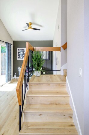 Una escalera con escalones de madera y barandal de metal negro que conduce a una habitación con ventilador de techo y planta en maceta.