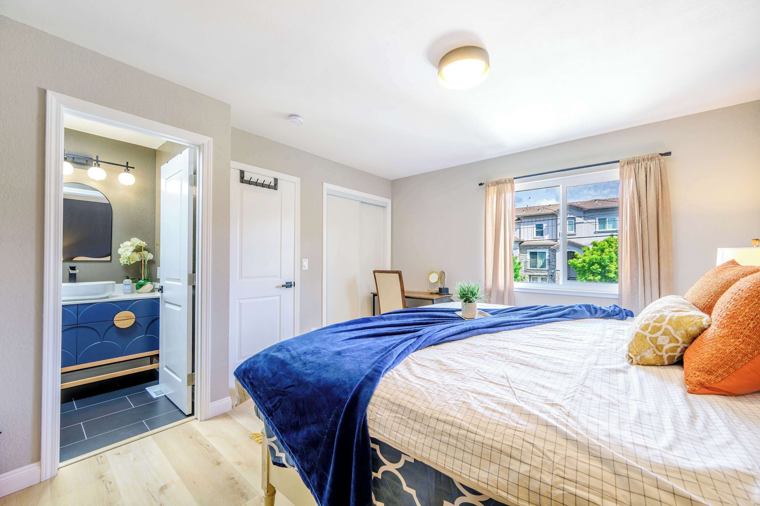 A bedroom with a large bed, a window with curtains, a bedside table, and a view of buildings outside.