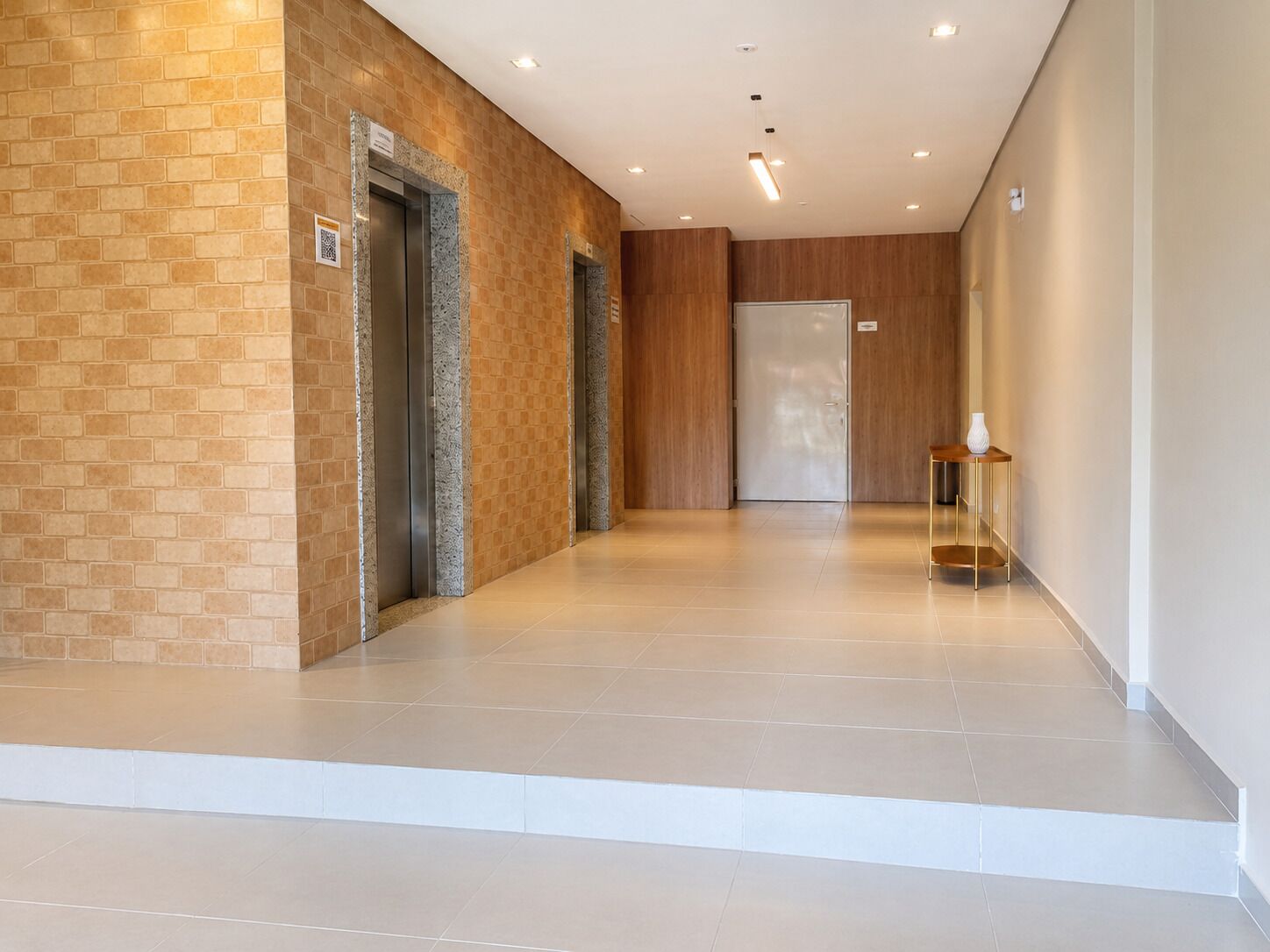 Corredor moderno com dois elevadores, uma porta de madeira e uma pequena mesa com um vaso.