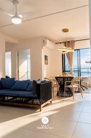 A modern living room with a sofa, dining table, and chairs, a ceiling fan, and a large window with a view.