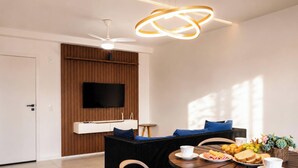 Ein modernes Wohnzimmer mit Flachbildfernseher an einer Wand aus Holzpaneelen, Deckenventilator und Essbereich mit gedecktem Tisch.