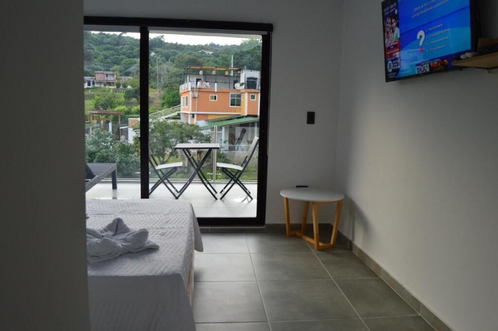 A hotel room with a bed, a small table, a chair, a TV, and a balcony with a view of buildings and greenery.
