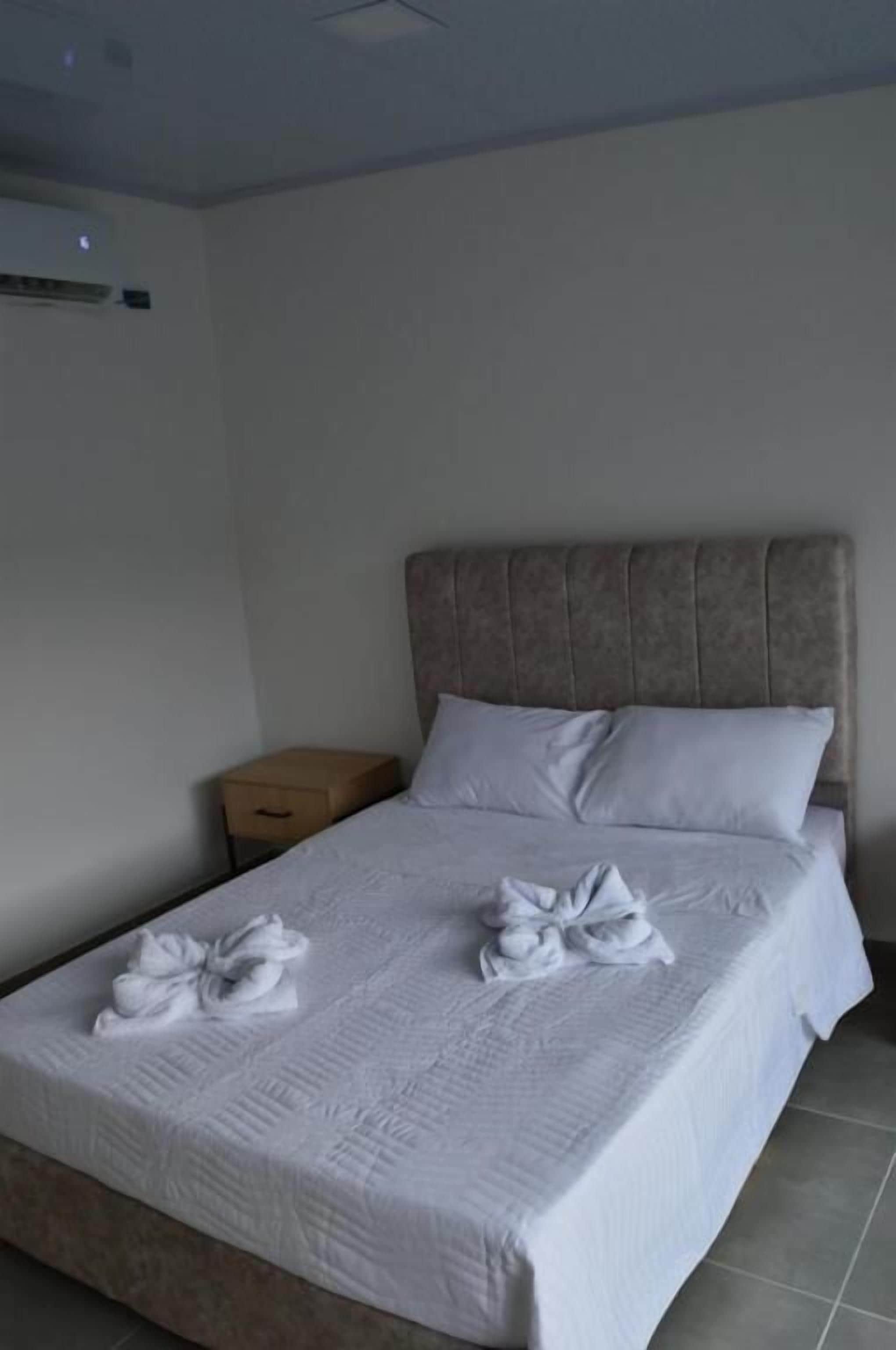 Een net opgemaakt bed met witte lakens en handdoeken, een houten nachtkastje en een wandairconditioning.