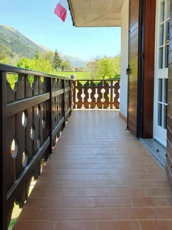 Ein überdachter Balkon mit Fliesenboden, Holzgeländer mit runden Ausschnitten und Blick auf grüne Hügel im Hintergrund.