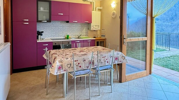 A kitchen with purple cabinets, a dining table with chairs, and a sliding glass door leading to a balcony. - Casa Via Vai - Casa in Scalve (Valle Richetti)