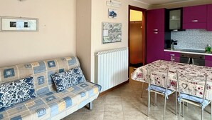 A living room with a sofa, a dining table with chairs, a kitchen area with purple cabinets, and a radiator. - Casa Via Vai - Casa in Scalve (Valle Richetti)
