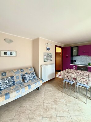 Een woonkamer met een bank, een eettafel met stoelen, een keuken met paarse kastjes en een radiator.