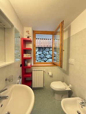 Un bagno con due lavandini, un water, uno scaffale rosso, una finestra con vista sulle rocce e uno specchio.