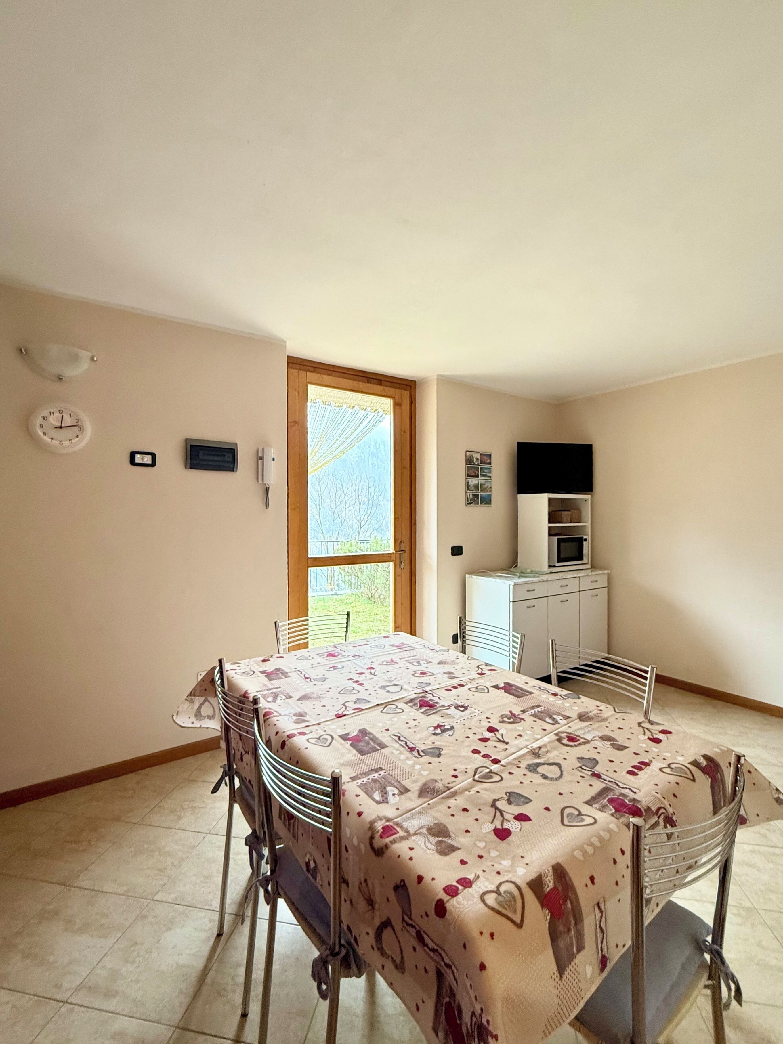 A small, well-lit room with a dining table, chairs, a kitchenette, and a door leading to a garden.
