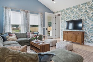 A living room with a sofa set, a coffee table, a TV on the wall, and a patterned wallpaper. - Luxury Coastal Family Retreat-Fire Pit, Game Room, Walk to Beach, Kid Friendly! (Gulfport)