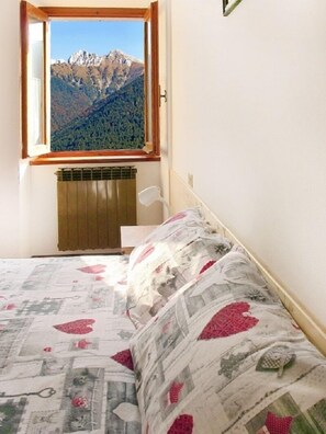 Une chambre avec vue sur la montagne, un radiateur, un lit recouvert d’une couvre-lit à motifs floraux et une fenêtre avec un cadre en bois.