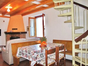 Une pièce chaleureuse avec une cheminée, une table à manger recouverte d’une nappe à motifs, un banc en bois et un escalier en colimaçon.