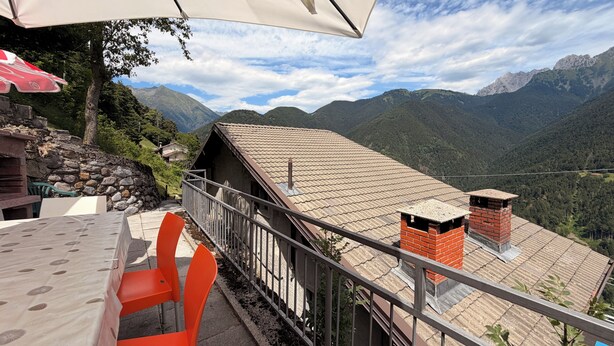 Terraza con vista a la montaña, una mesa, sillas, un muro de piedra y un edificio con tejado de baldosas.