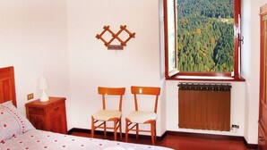 Un dormitorio con una cama, dos sillas, una mesita de noche, una lámpara, un radiador y una ventana con vistas a un bosque.