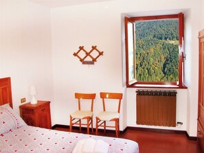 Une chambre à coucher comprenant un lit, deux chaises, une table de chevet, une lampe, un radiateur et une fenêtre donnant sur une forêt.