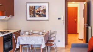 A dining area with a table set for four, a kitchen with a stove and oven, a framed picture of a canal, and a door leading to another room. - Quater Pis - Casa in Scalve (Colere)