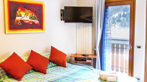 A hotel room with a bed, pillows, a TV, a small table, and a view of a balcony. - Quater Pis - Casa in Scalve (Colere)
