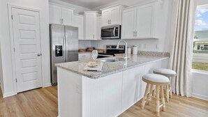 Cocina moderna con armarios blancos, encimera de granito, refrigerador de acero inoxidable, microondas y un lavamanos. Hay dos taburetes de madera y una ventana con cortinas.