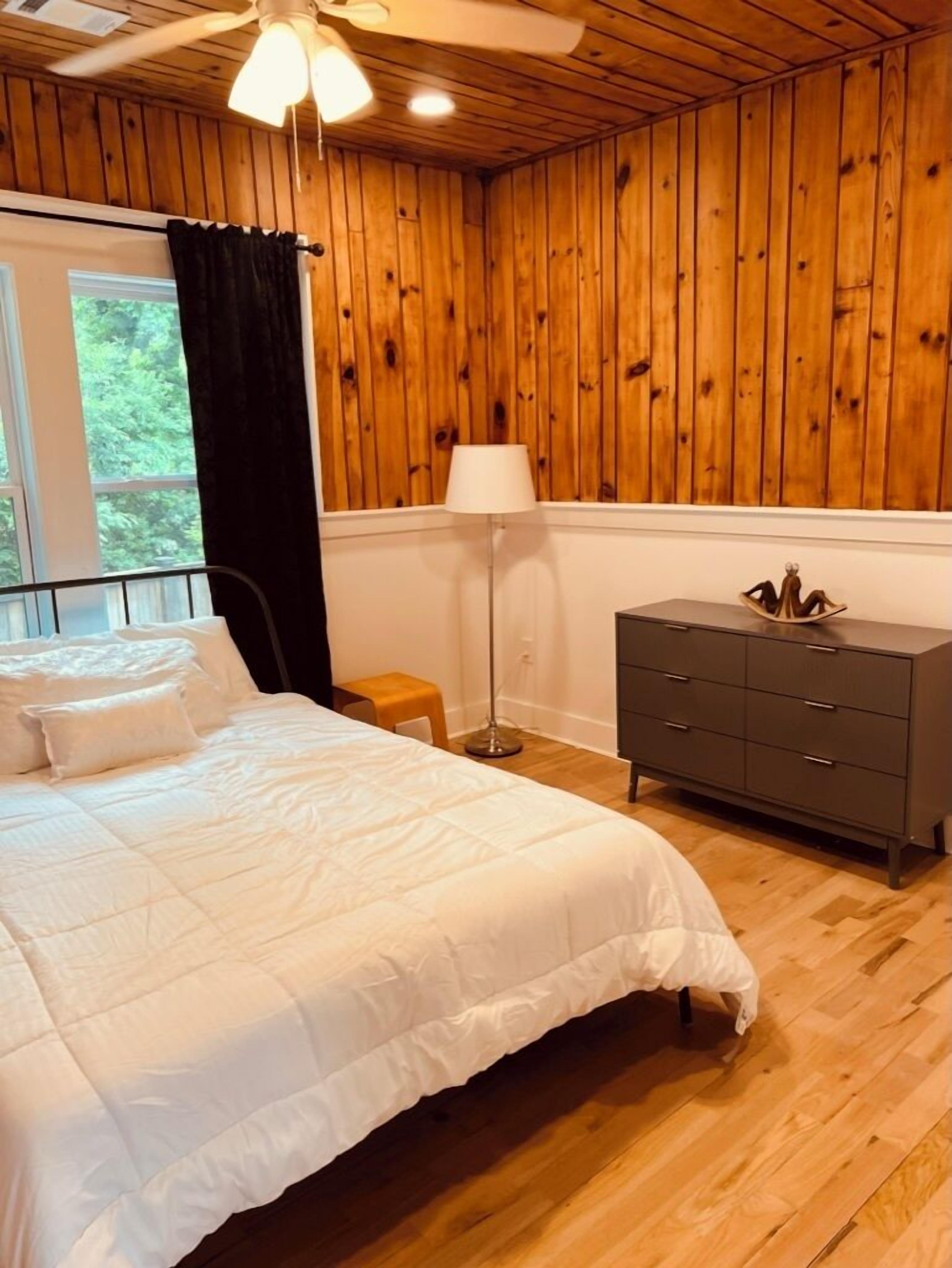 Une chambre avec un mur lambrissé en bois, un lit avec une literie blanche, une lampe sur pied et une commode.