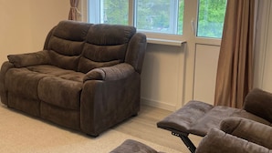 A room with a brown sofa, a window with curtains, and a view of greenery outside.