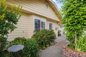 A beige house with a brick pathway leading to the entrance, surrounded by lush greenery and flowering plants.