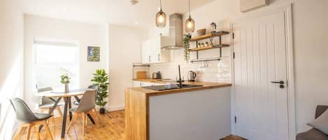 A modern kitchen with a central island, wooden flooring, pendant lighting, and a dining area with chairs.