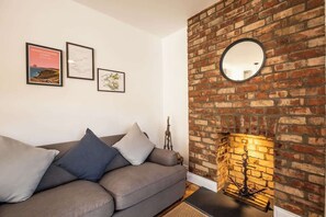 A living room with a brick fireplace, a sofa with cushions, and framed artwork on the wall. - Ty Llwyd Ceibwr - Modern apartment (Saint Dogmaels)