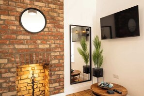 A room with a brick wall, a round mirror, a flat-screen TV, a wooden table with a bowl and remote, and a lit fireplace. - Ty Llwyd Ceibwr - Modern apartment (Saint Dogmaels)