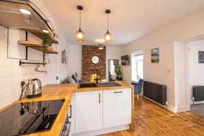 Une cuisine moderne avec un plan de travail en bois, des armoires blanches, un mur d’accent en briques et des luminaires suspendus.