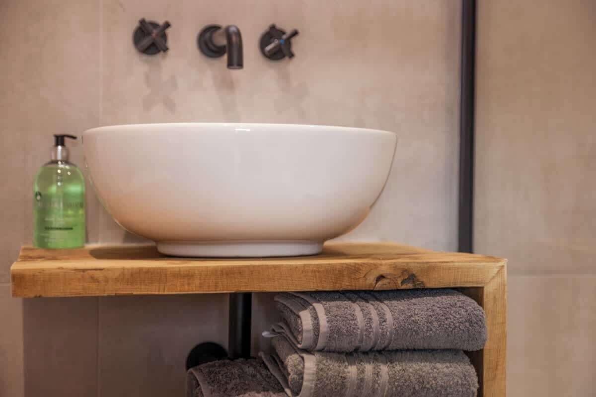 A bathroom with a large white basin on a wooden shelf, a green bottle of soap, and folded towels.