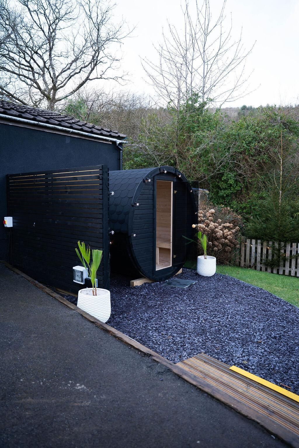 A building with a large cylindrical structure, a black exterior, and a wooden door. There are two potted plants in front.