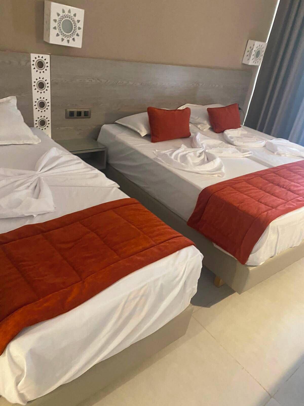 A hotel room with two beds, each with white linens and red accents, a nightstand, and wall-mounted decor.
