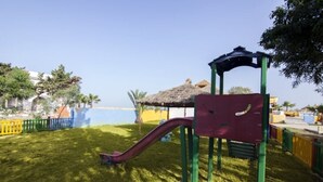 Un patio de juegos con un tobogán y estructuras para escalar, rodeado por una zona vallada con césped.