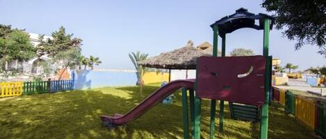 Un patio de juegos con un tobogán y estructuras para escalar, rodeado por una zona vallada con césped.