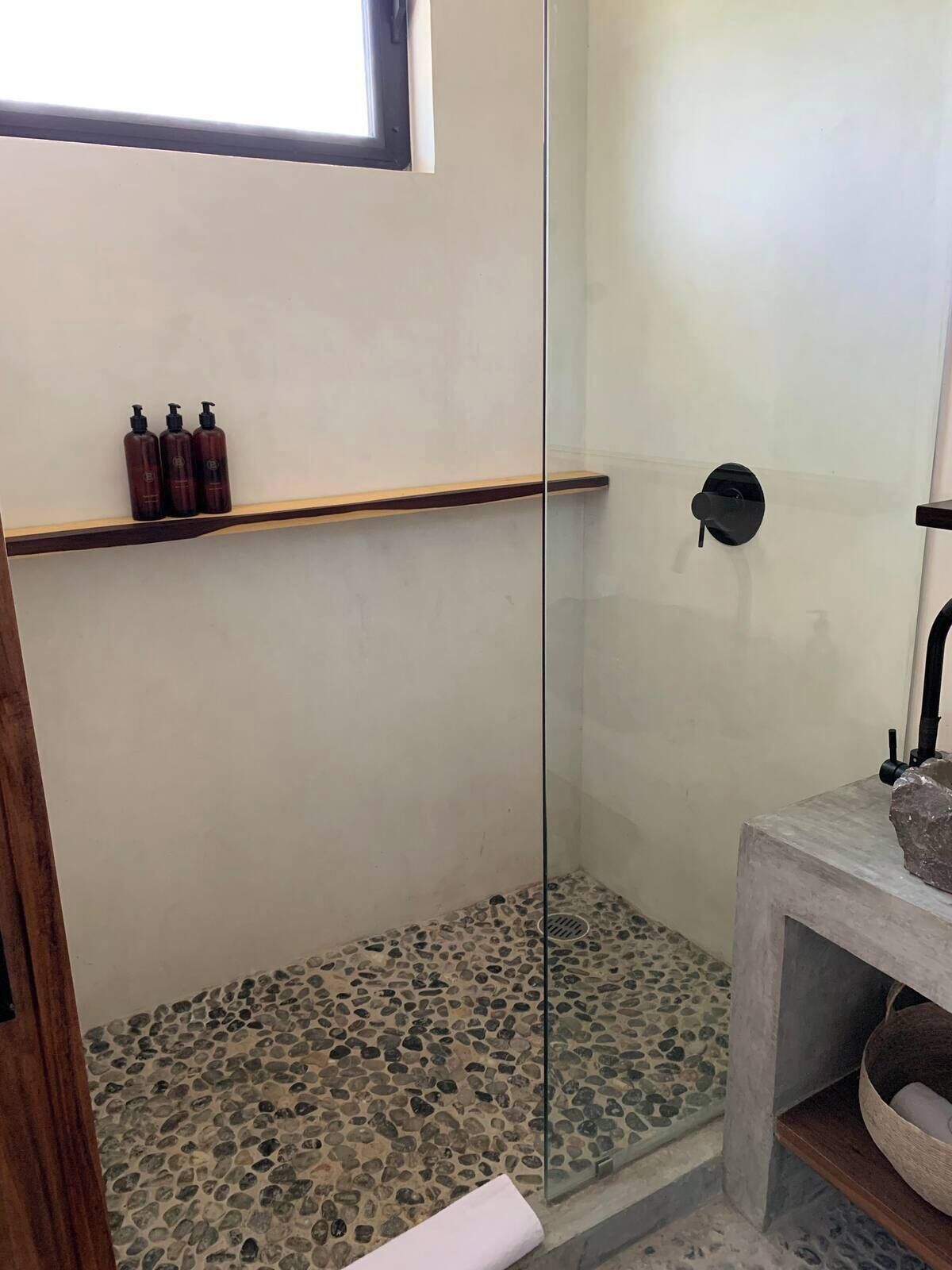 Une douche avec une porte en verre, une étagère en bois où sont rangés les articles de toilette et un sol imitant la pierre.