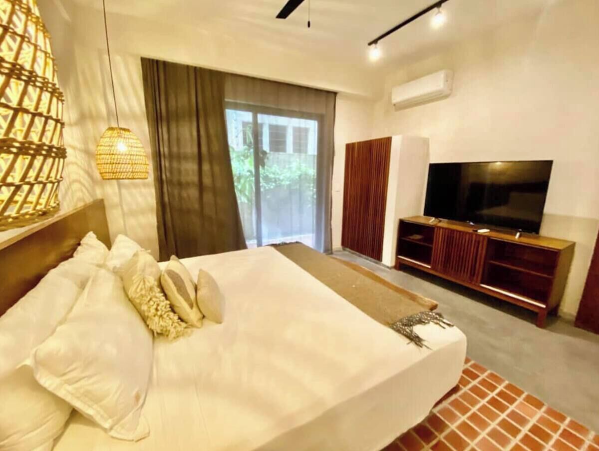 A modern hotel room with a large bed, a TV, a wooden entertainment unit, and a view of greenery through the window.