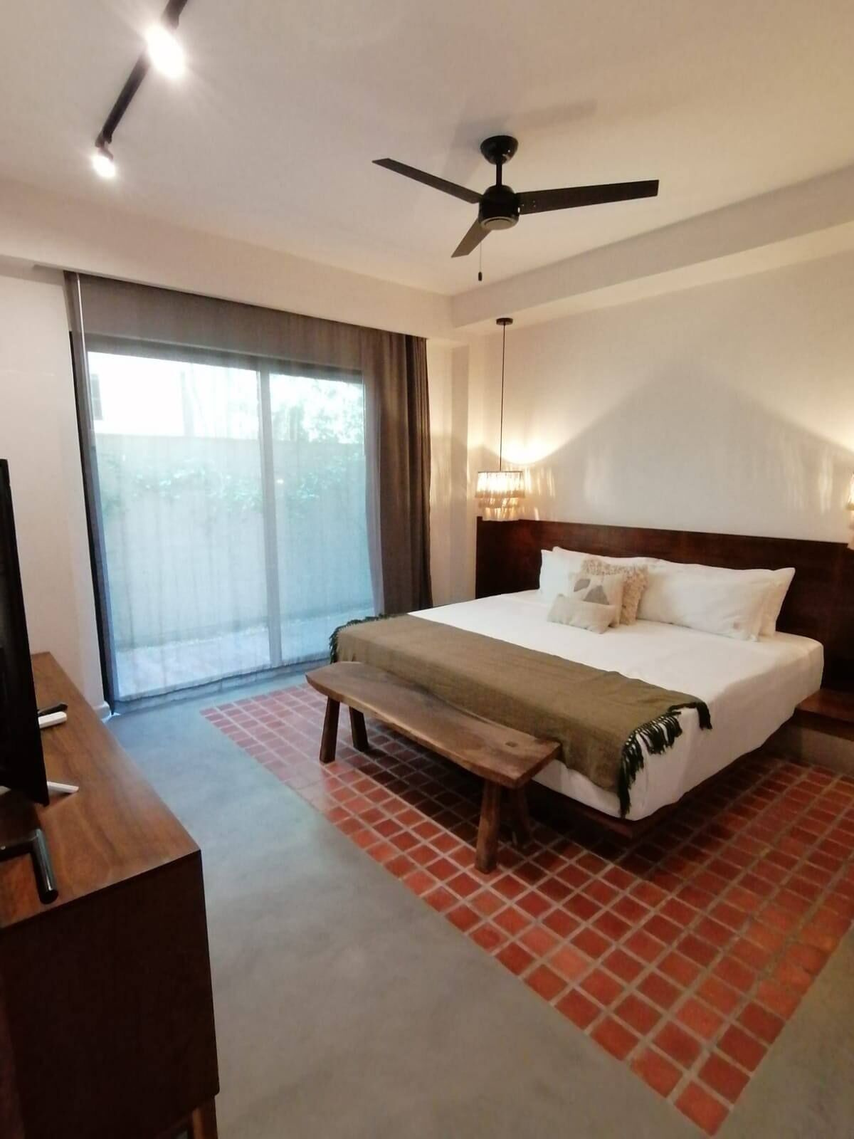 Habitación de hotel con cama, banca, televisor, ventilador de techo y un amplio ventanal.