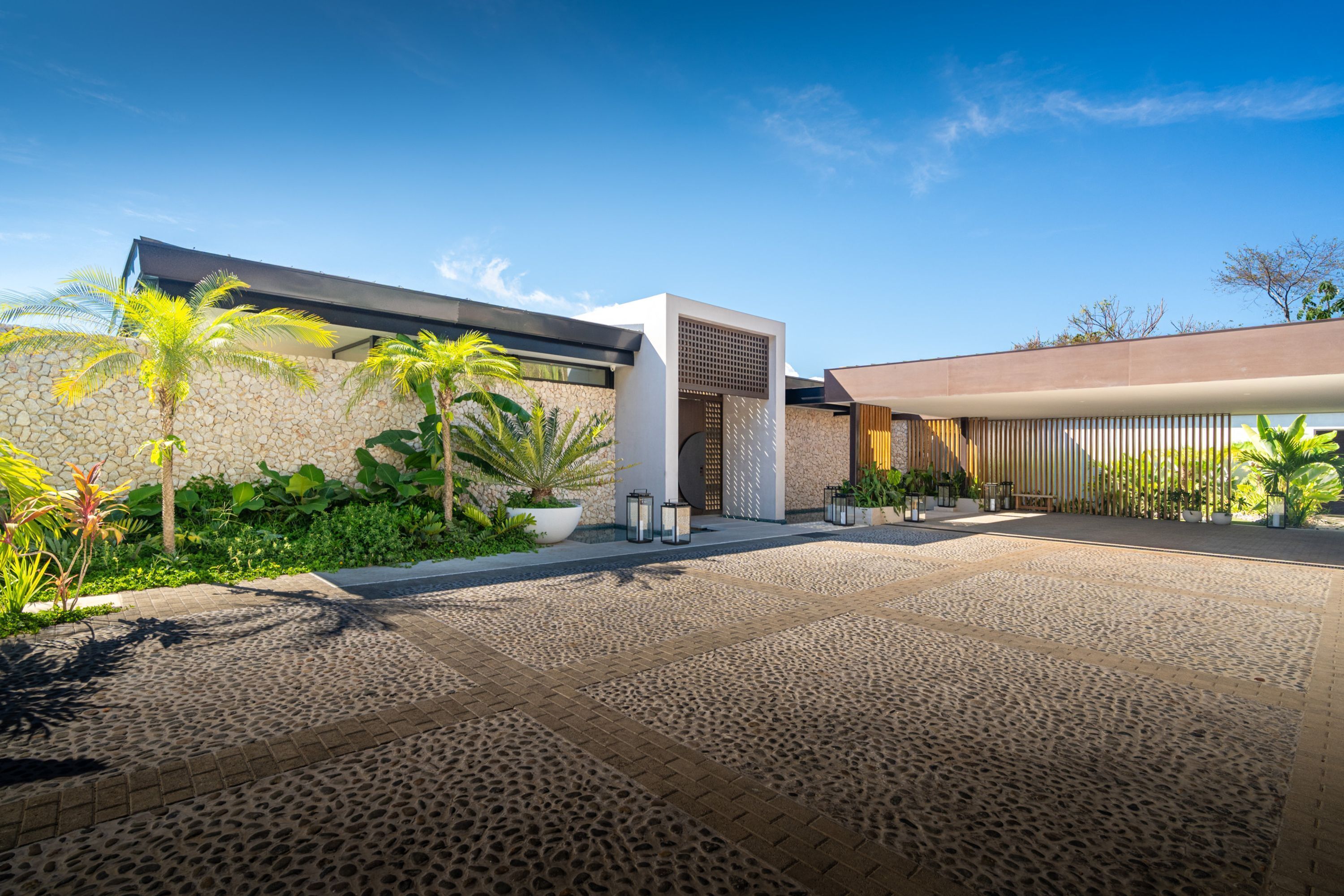 A modern building with a stone facade, a paved courtyard, and tropical plants.