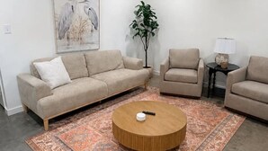 Een woonkamer met een hoekbank, een ronde salontafel, een kamerkleed met patroon, een vloerlamp en een aan de muur hangend schilderij.