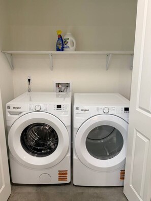 Two Whirlpool front-loading washing machines in a laundry room with a shelf above containing cleaning supplies. - Decatur Inn•3 KINGS•Lux Amenities•Fast Wi-Fi (Lafayette)