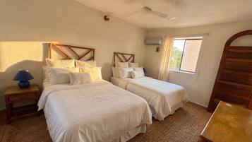 A room with two beds, a ceiling fan, a nightstand with a lamp, a window with curtains, and a wooden door.