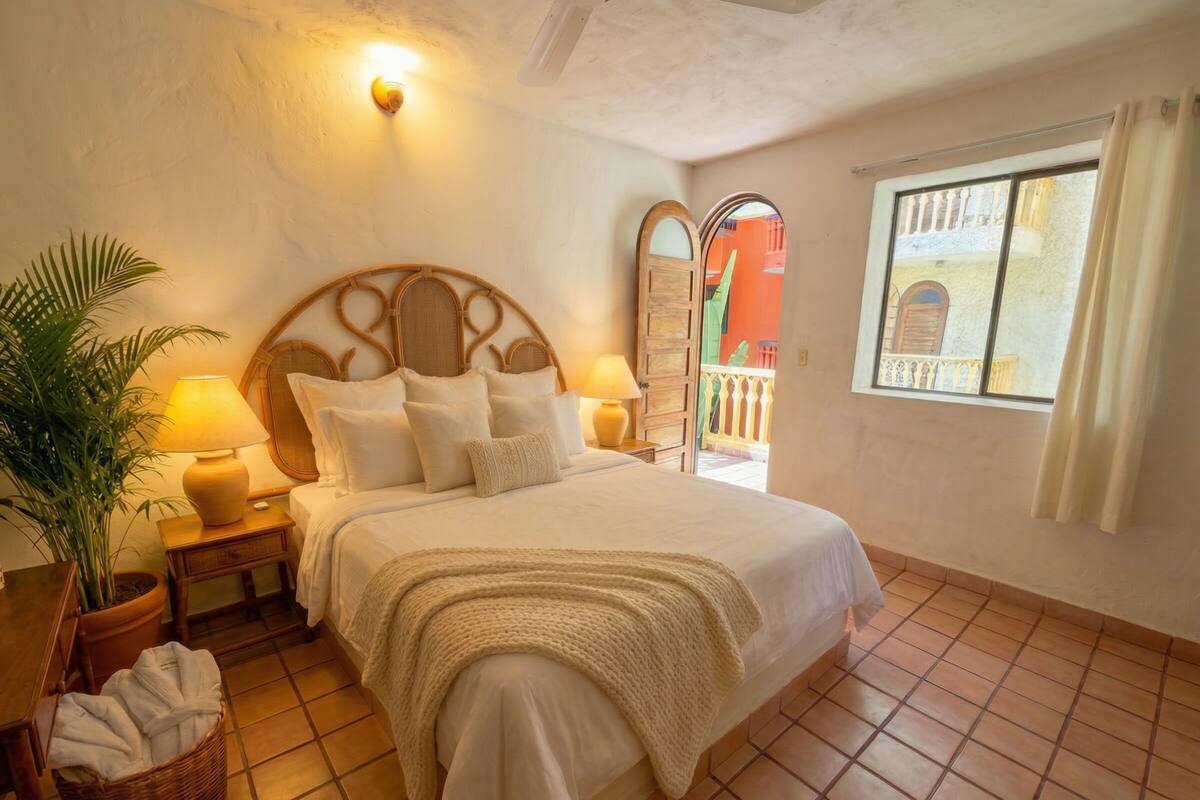 A bedroom with a bed, bedside lamps, a potted plant, a nightstand, and a window with curtains.