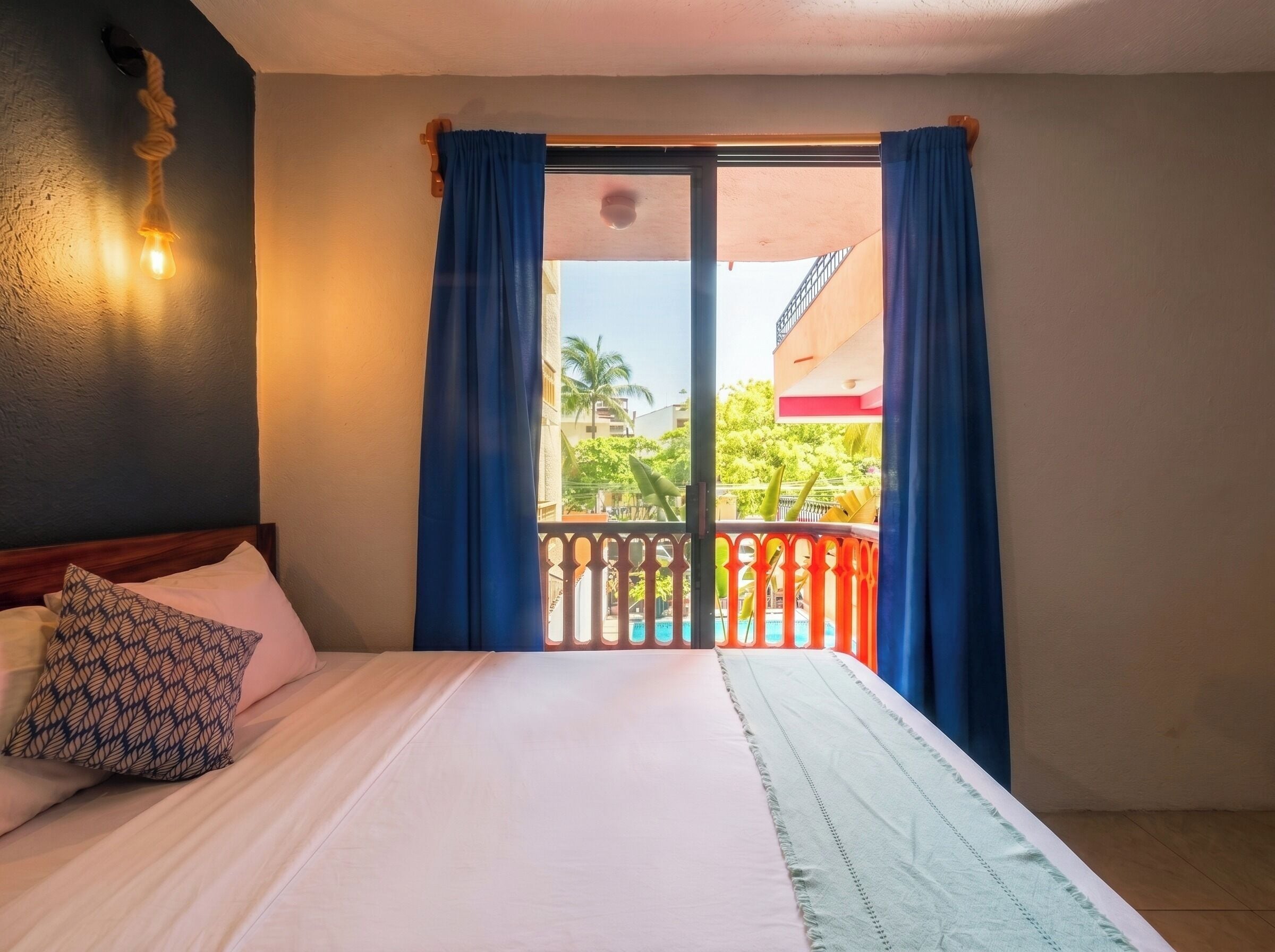 Habitación de hotel con cama, ventana con cortinas azules, balcón con barandilla y lámpara fijada a la pared.