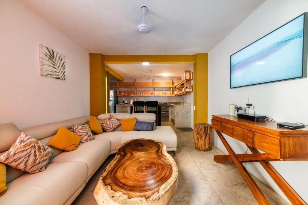 A living room with a sofa, coffee table, and a TV. There is a kitchen visible in the background.