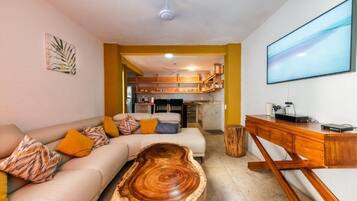 A living room with a sofa, coffee table, and a TV. There is a kitchen visible in the background.